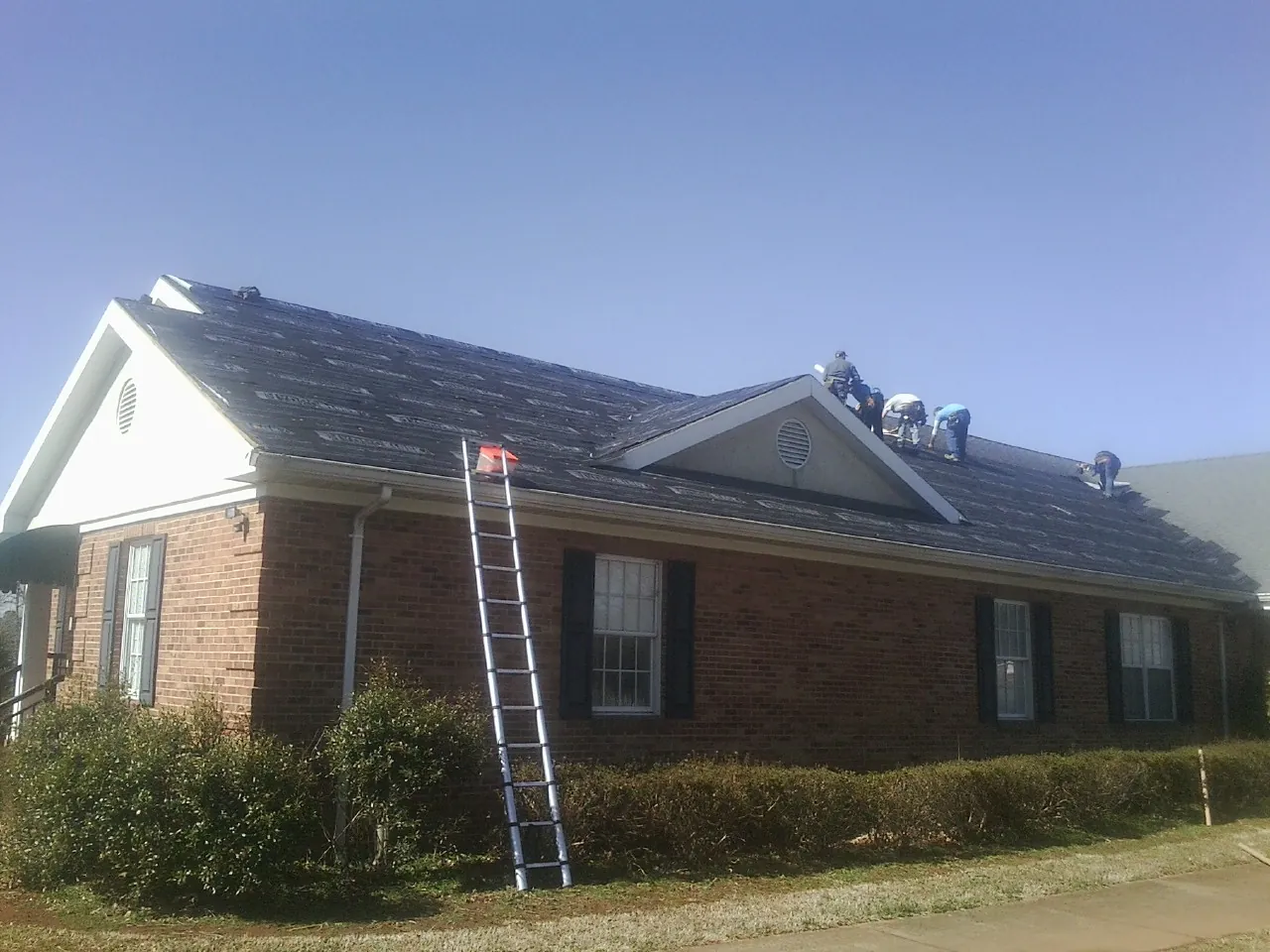 Crew tearing shingles off a brick ranch roof with a ladder set — Emergency Roof Repair work by our crew in Poca
