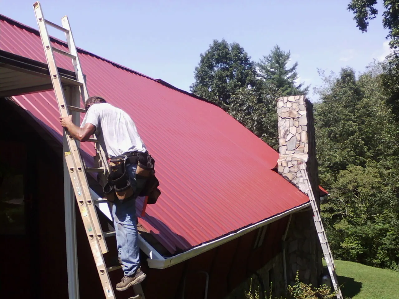Roofer on a ladder beside a red metal roof and stone chimney — Soffit & Fascia Repair by Roofing Brothers in Poca